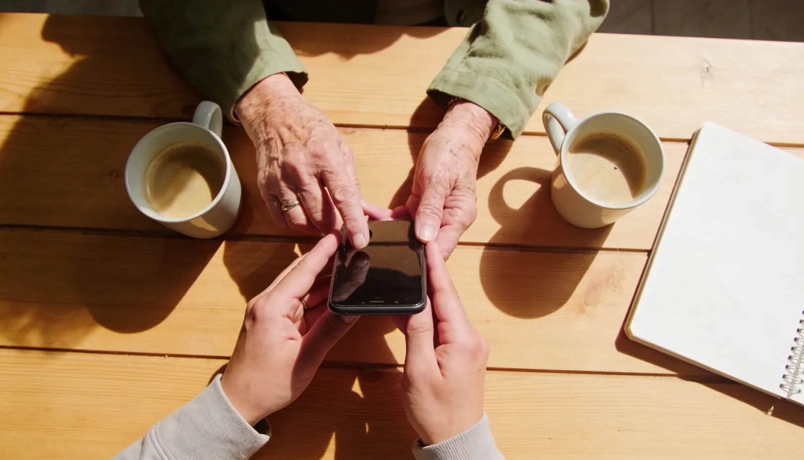 Vista zenital d'una taula de fusta amb mans de dues generacions al voltant d'un telèfon mòbil i dos cafés